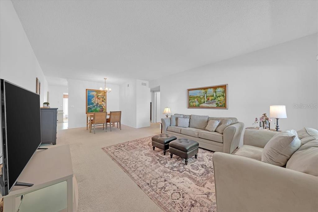 A living room with beige carpet, a sofa, two ottomans, and an adjacent dining area — 2401 FRANCISCAN DRIVE UNIT 41, CLEARWATER, FL