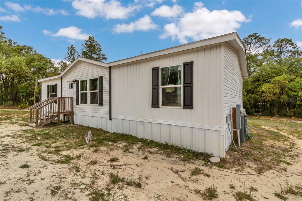 A white manufactured home with black shutters sits on a sandy lot surrounded by trees — 5021 S ADVENTURE POINT, HOMOSASSA SPRINGS, FL