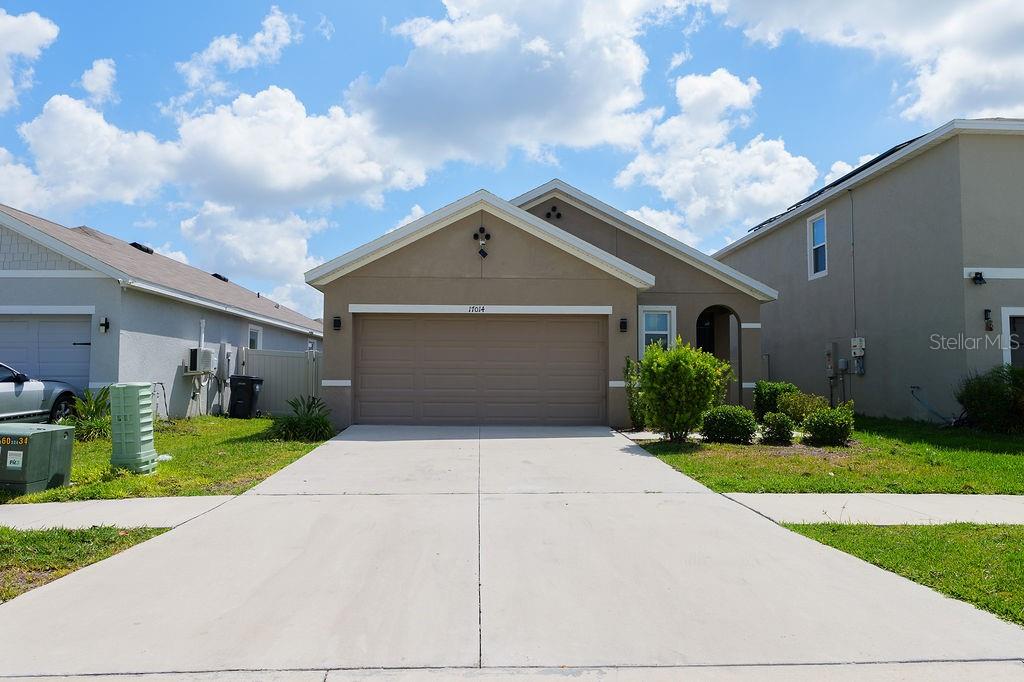 A tan single-story house features a two-car garage and a concrete driveway under a blue sky — 17014 BLISTER WING DRIVE, WIMAUMA, FL