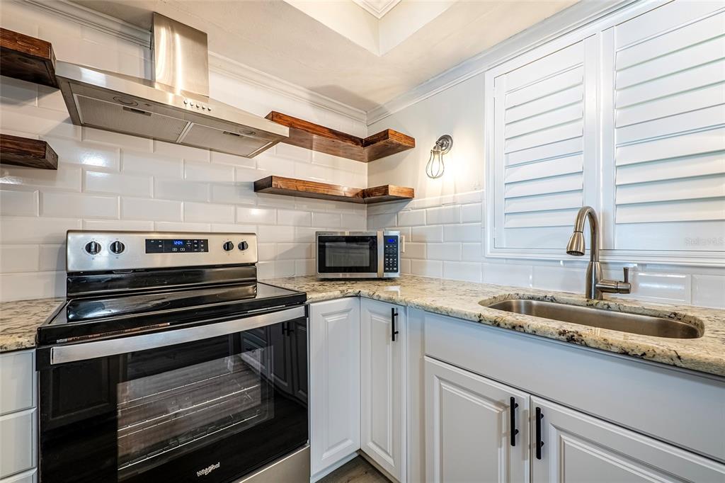 A kitchen features white cabinets, subway tile backsplash, granite countertops, and open wood shelving — 620 EDGEWATER DRIVES UNIT 103, DUNEDIN, FL
