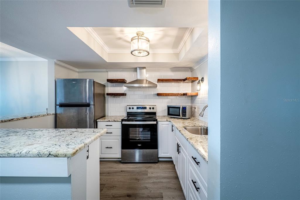 A kitchen features white cabinets, granite countertops, stainless steel appliances, and floating wooden shelves — 620 EDGEWATER DRIVES UNIT 103, DUNEDIN, FL