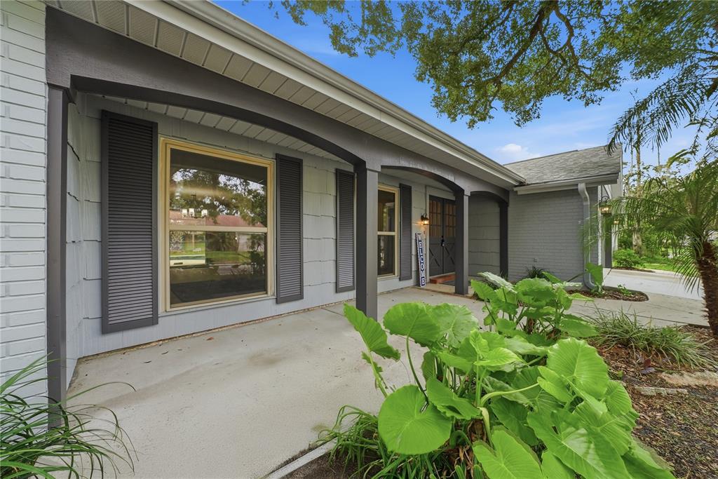 A covered front porch with gray siding, dark shutters, and large green leafy plants — 12605 RIVER MILL DRIVE, HUDSON, FL
