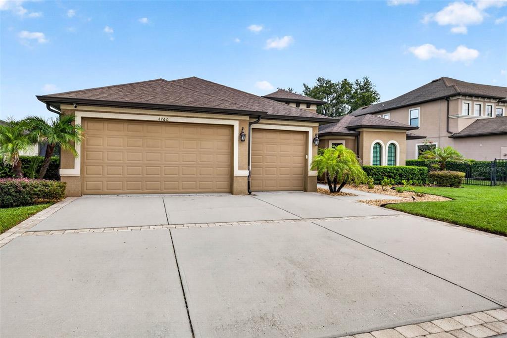 A tan house with a three-car garage and a concrete driveway under a blue sky — 4760 LAGO VISTA CIRCLE, LAND O LAKES, FL