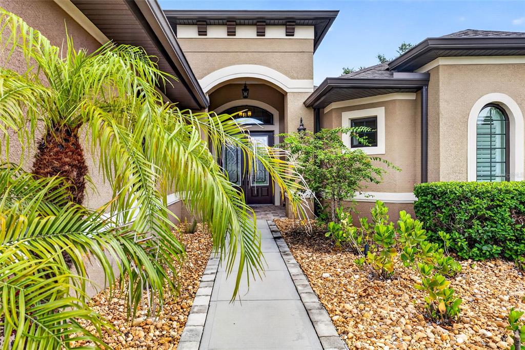 A concrete walkway leads to the arched entryway of a tan stucco house with landscaping — 4760 LAGO VISTA CIRCLE, LAND O LAKES, FL