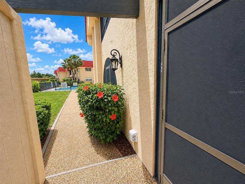 A paved walkway leads past a hibiscus bush toward a building with a screen door — 211 RUBENS DRIVE UNIT E, NOKOMIS, FL