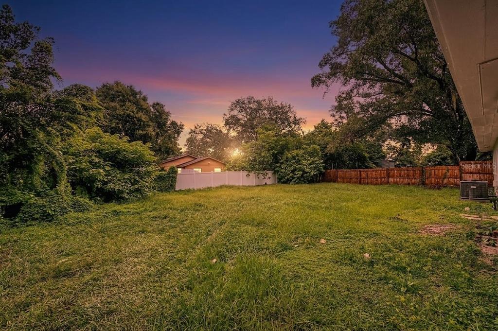 A large, grassy backyard features a white vinyl fence, mature trees, and a sunset sky — 810 E 120TH AVENUE, TAMPA, FL
