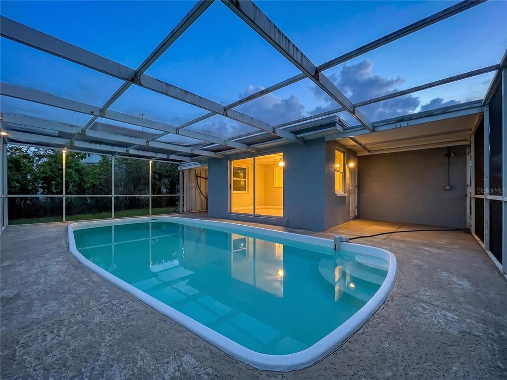 A swimming pool sits inside a screened-in patio area next to a gray house — 7513 HATTERAS DRIVE, HUDSON, FL
