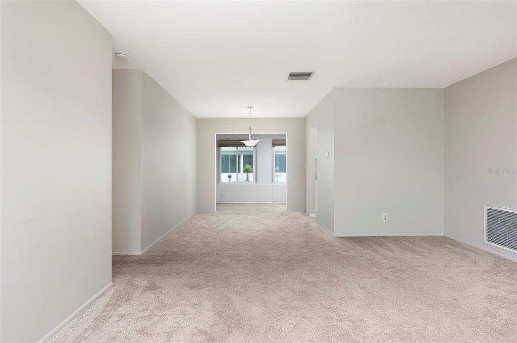 An empty room with beige carpeting and light-colored walls leads to a sunlit dining area — 3710 98TH AVENUE N, PINELLAS PARK, FL