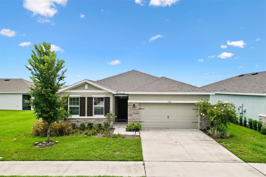 A single-story suburban house with a tan stucco exterior, stone accents, and a two-car garage — 7926 PENROSE PLACE, WILDWOOD, FL