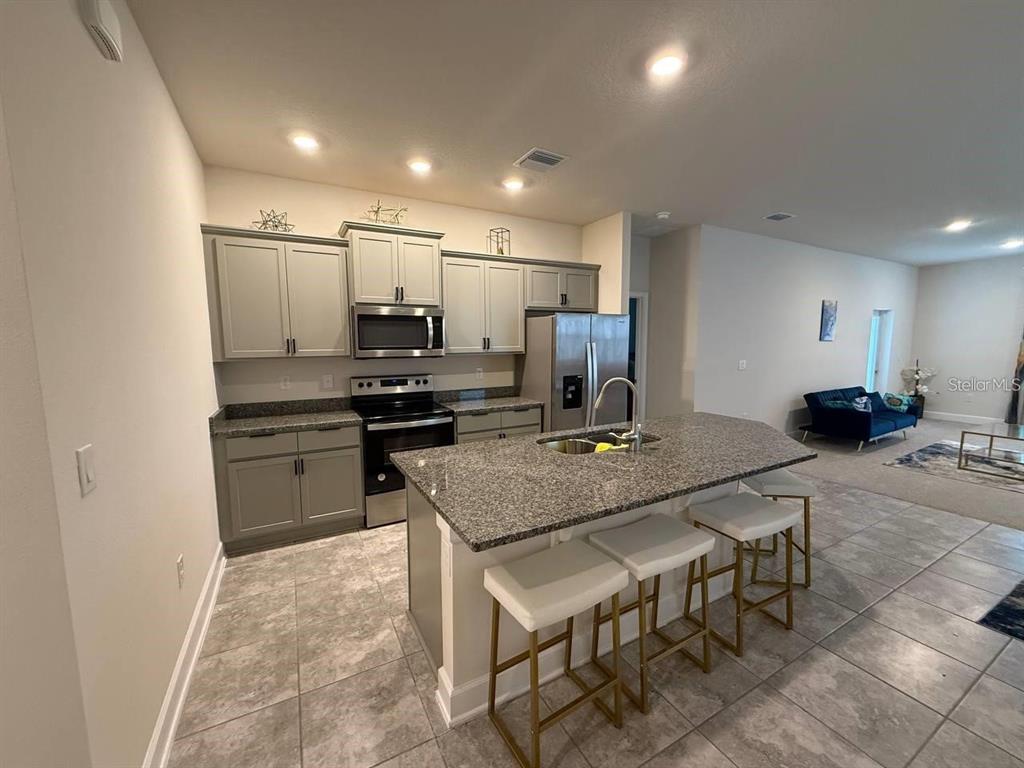 A kitchen features gray cabinets, granite countertops, a center island with bar stools, and tile flooring — 3828 SAGEFIELD DRIVE, HARMONY, FL