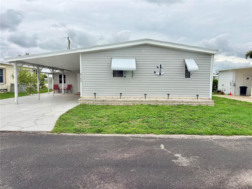 A single-story manufactured home with light gray siding, a carport, and a brick-patterned base — 3163 INDIAN VILLAGE LANE, NORTH FORT MYERS, FL