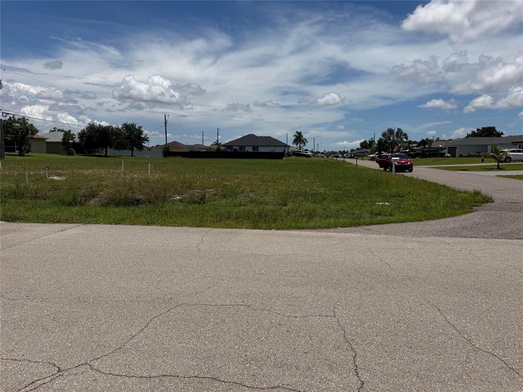 A vacant, grassy residential lot sits at a street corner under a cloudy blue sky — 2829 NE 5TH AVENUE, CAPE CORAL, FL