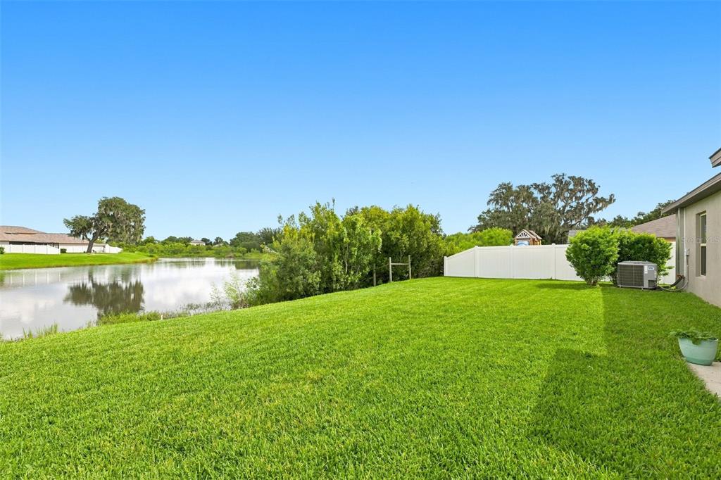 A grassy backyard features a pond, a white vinyl fence, and a house exterior — 39745 HARVEST CREEK LANE, ZEPHYRHILLS, FL