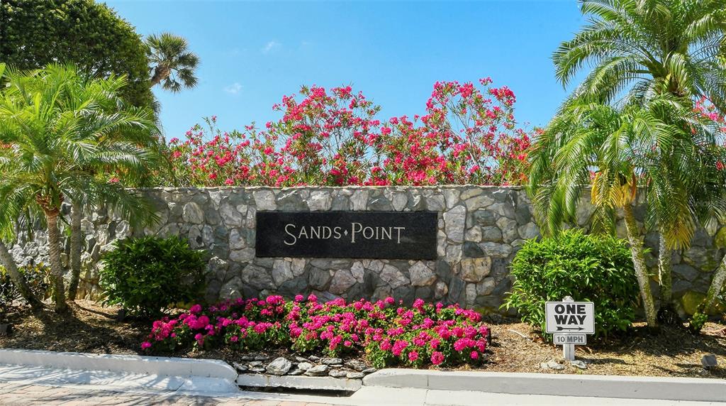 A stone wall with a Sands Point sign stands behind pink flowers and palm trees — 100 SANDS POINT ROAD UNIT 324, LONGBOAT KEY, FL