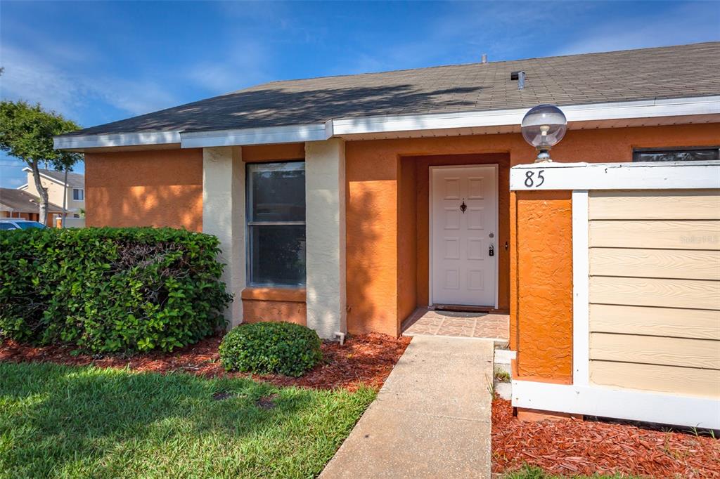 The exterior of a single-story home features orange stucco walls, a white door, and landscaping — 85 SILVER PARK CIRCLE, KISSIMMEE, FL