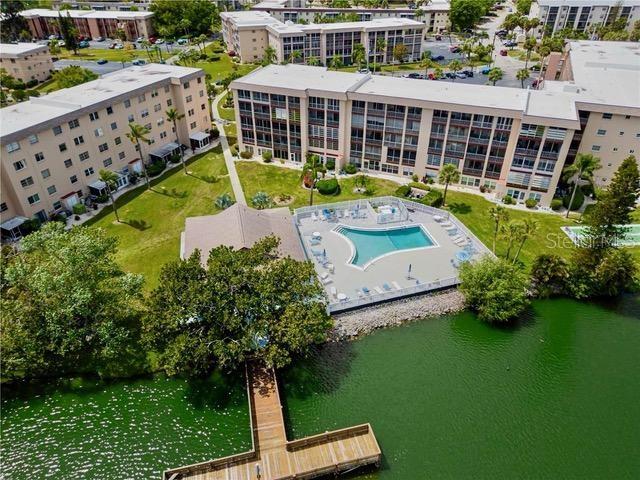 An aerial view shows apartment buildings, a swimming pool, and a wooden dock on a lake — 4145 HERON WAY UNIT E-208, BRADENTON, FL