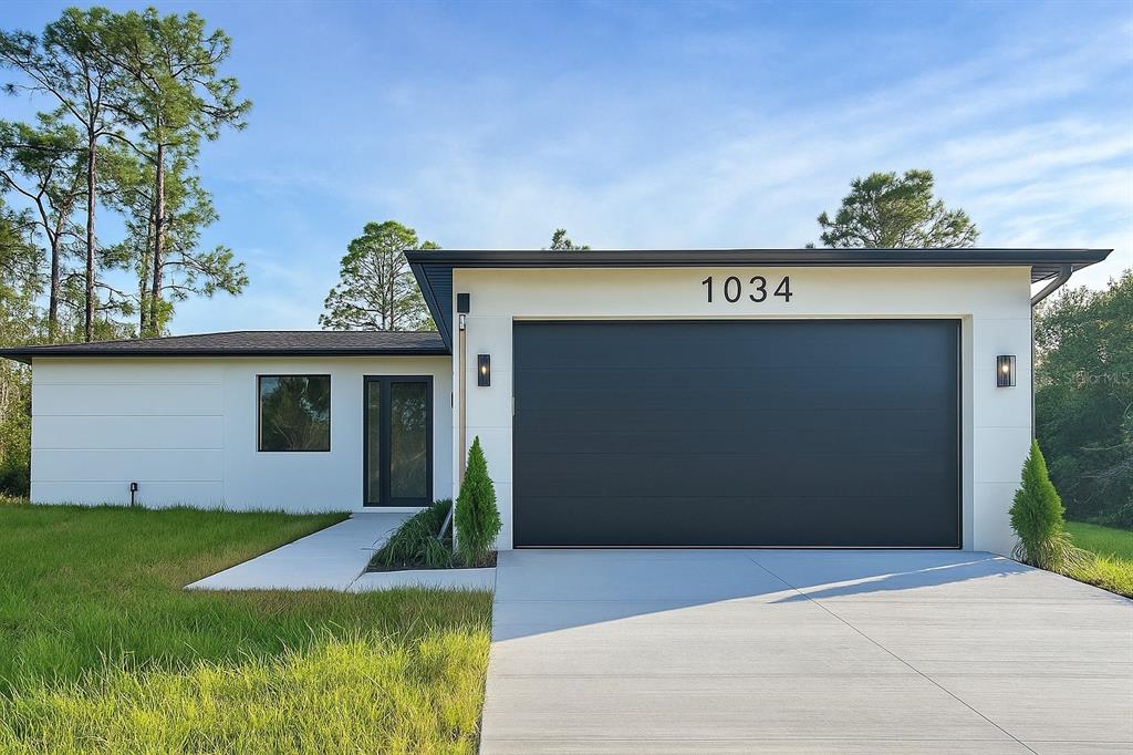 A modern single-story house features a white exterior, a dark garage door, and a walkway — 1034 ALCALDE STREET E, LEHIGH ACRES, FL
