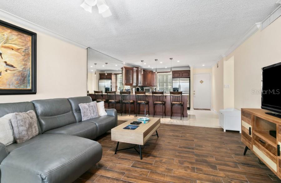 A living room with wood-look tile flooring features a gray sectional sofa and a kitchen — 605 SUTTON PLACE UNIT 102, LONGBOAT KEY, FL