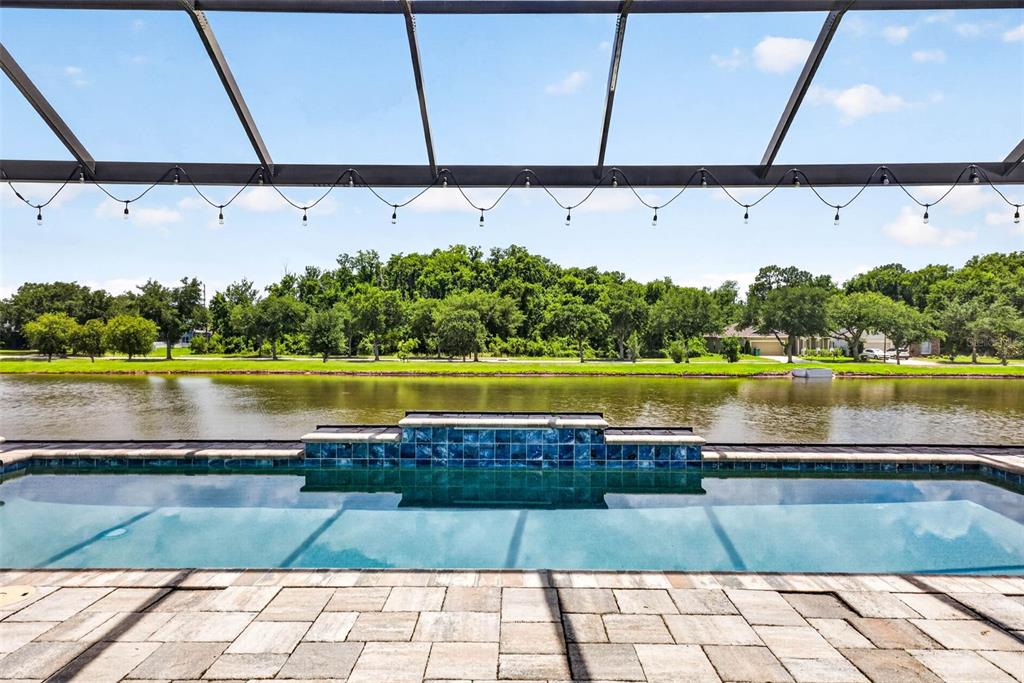 A swimming pool with a blue tile water feature overlooks a lake and green trees — 6946 TALAMORE DRIVE, WESLEY CHAPEL, FL