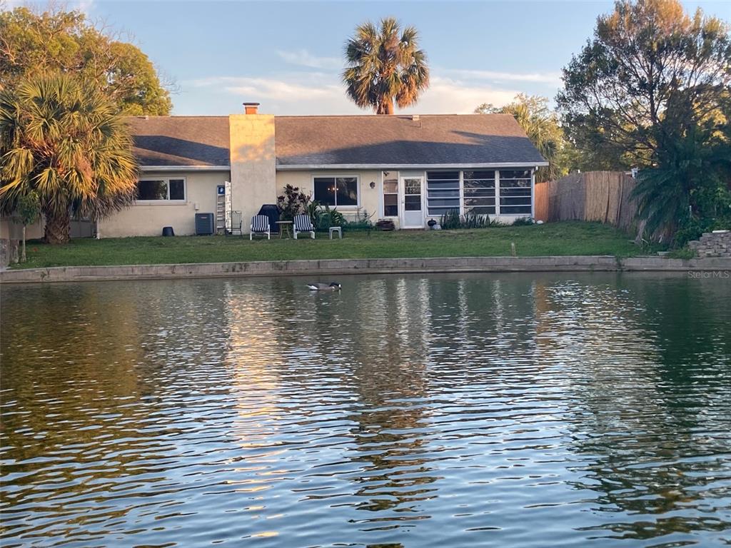 A single-story house with a chimney and a screened porch sits beside a pond — 7561 HOLLY LAKE LANE, NEW PORT RICHEY, FL