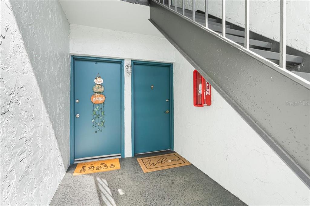 Two teal doors with welcome mats sit beneath an outdoor staircase next to a fire extinguisher — 11401 3RD STREET N UNIT 2, ST PETERSBURG, FL