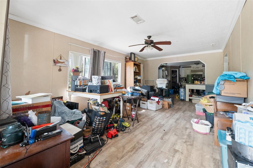 A cluttered living room with wood-look flooring, a ceiling fan, and an open kitchen beyond — 17221 NICKS DRIVE, SPRING HILL, FL