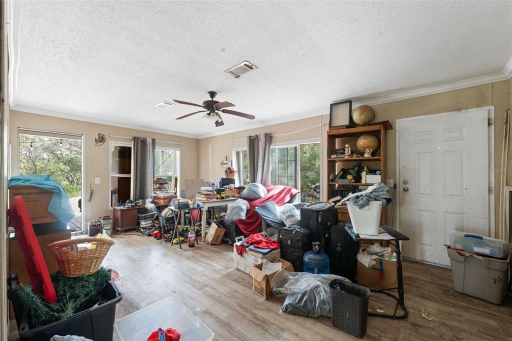A cluttered room with light wood-look flooring, a ceiling fan, and a white exterior door — 17221 NICKS DRIVE, SPRING HILL, FL