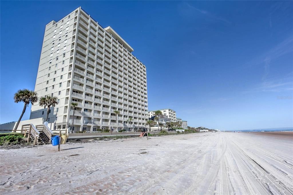 A tall, white beachfront condominium building stands next to a sandy beach under a clear sky — 89 S ATLANTIC AVENUE UNIT 204, ORMOND BEACH, FL