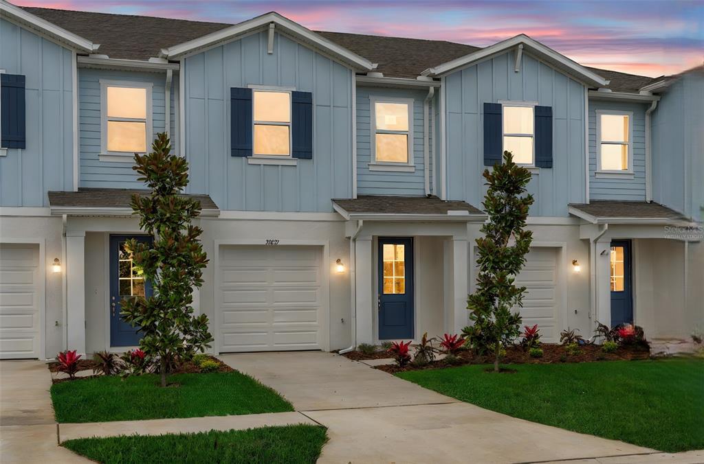 A row of light blue townhouses features white garage doors, blue front doors, and landscaped yards — 31789 KESTREL CALLING DRIVE, SAN ANTONIO, FL