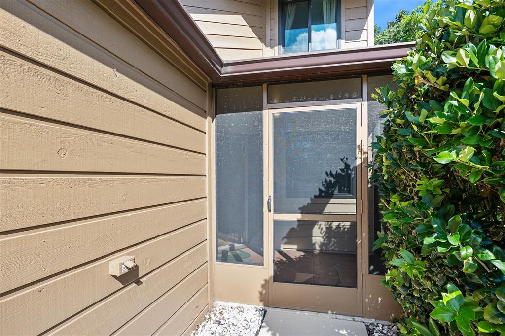 A tan-sided exterior wall meets a screened-in porch entrance next to a large green bush — 99 OLD BARN TRAIL, ORMOND BEACH, FL