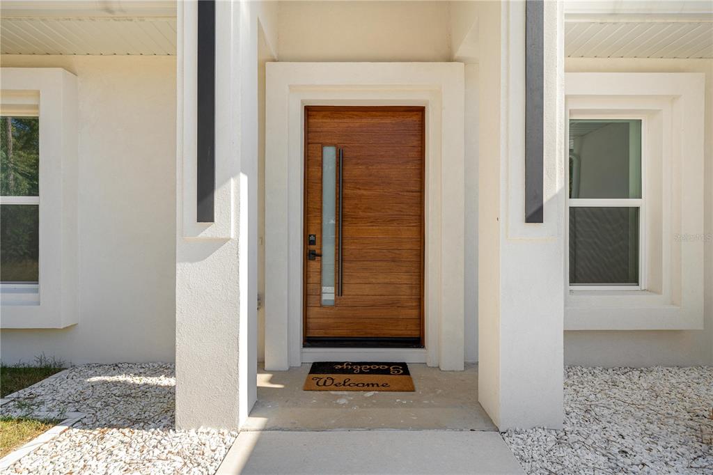 A modern wooden front door with a vertical glass panel sits between two white pillars — 6877 SW 131ST CIRCLE, OCALA, FL