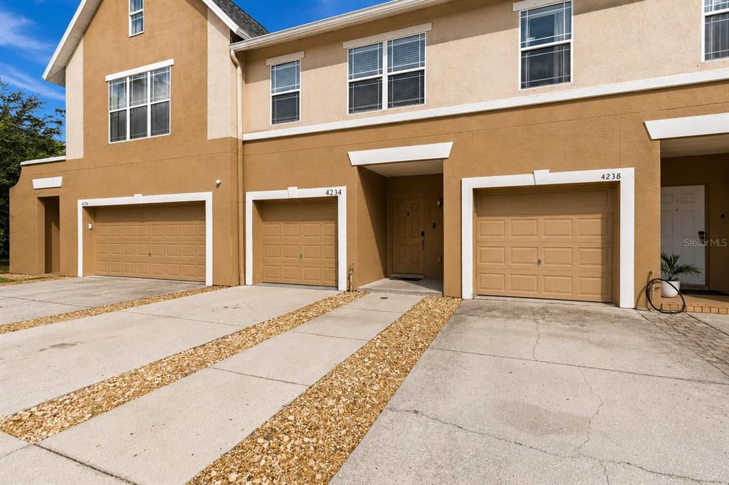 The exterior of a multi-unit townhouse features tan stucco walls, three garage doors, and driveways — 4234 TYLER CIRCLE N UNIT #102B, ST PETERSBURG, FL