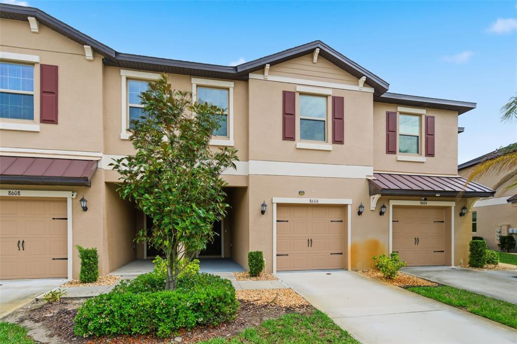 A row of three tan townhouses with attached garages and brown shutters under a blue sky — 8606 REEDVILLE STREET, NEW PORT RICHEY, FL