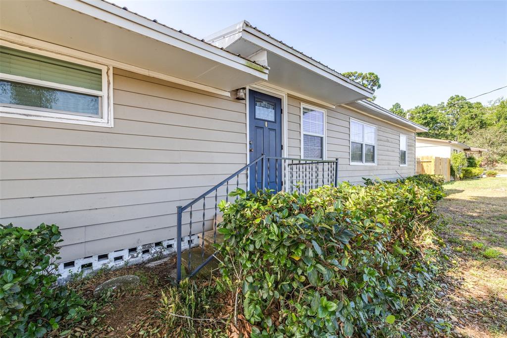 A beige house exterior features a blue front door, metal railing, and overgrown landscaping — 3935 CLUBHOUSE ROAD, HIGHLAND CITY, FL