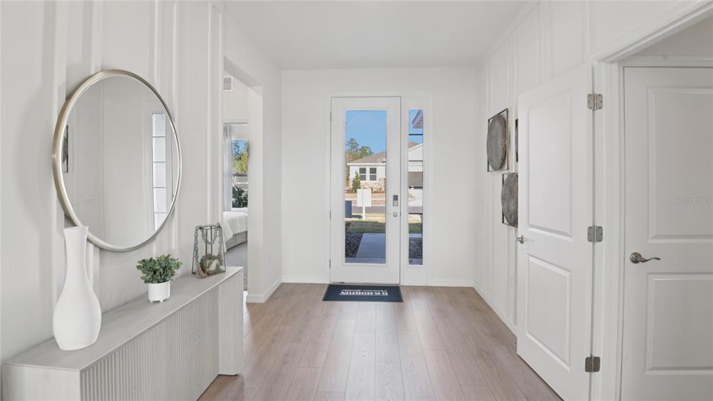 A bright entryway features light wood flooring, white board-and-batten walls, and a round wall mirror — 3049 CANOPY OAK BOULEVARD, WILDWOOD, FL