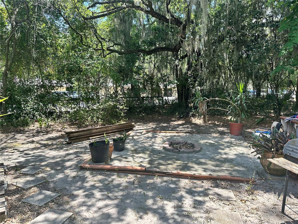 A backyard features a fire pit, potted plants, stacked lumber, and mature trees with moss — 919 NW 23RD AVENUE, GAINESVILLE, FL