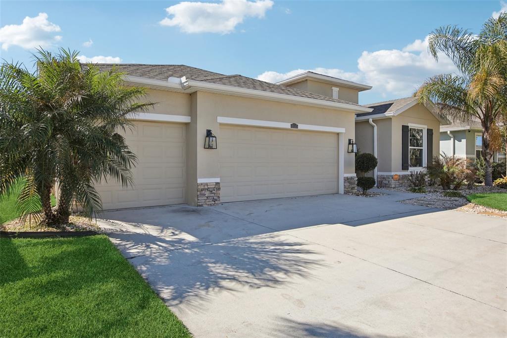 A beige house with a three-car garage, stone accents, and palm trees under a blue sky — 10747 LAXER CAY LOOP, SAN ANTONIO, FL