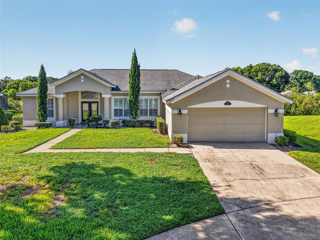 A single-story suburban house with a beige stucco exterior, a two-car garage, and manicured landscaping — 6022 FALCONBRIDGE PLACE, MOUNT DORA, FL