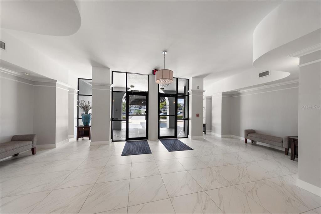 A bright lobby features white marble-look tile floors, two glass entry doors, and gray benches — 14820 RUE DE BAYONNE UNIT 505, CLEARWATER, FL