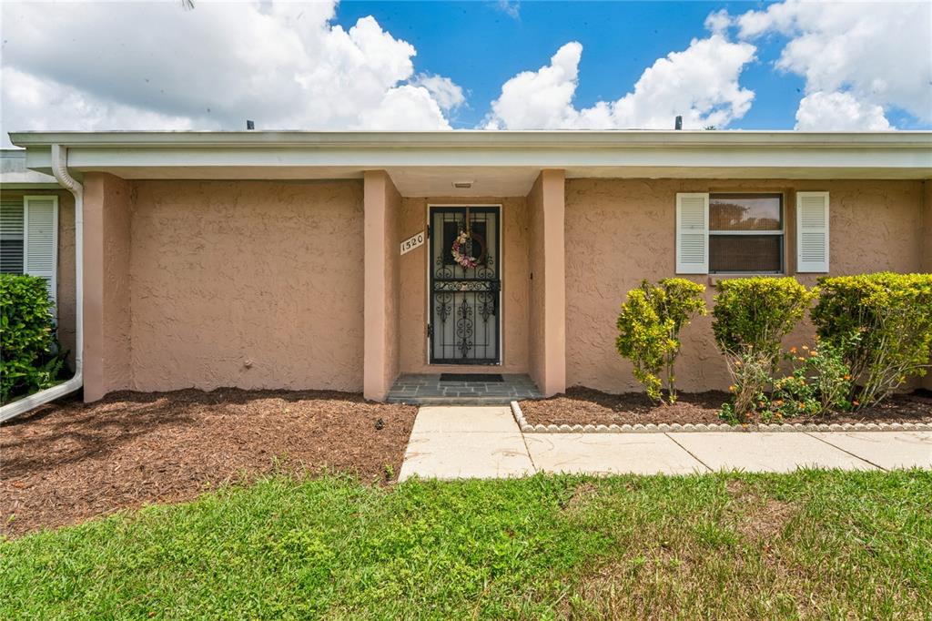 The exterior of a single-story house features a tan stucco facade, a front door, and landscaping — 1520 PALMER COURT, PALM HARBOR, FL