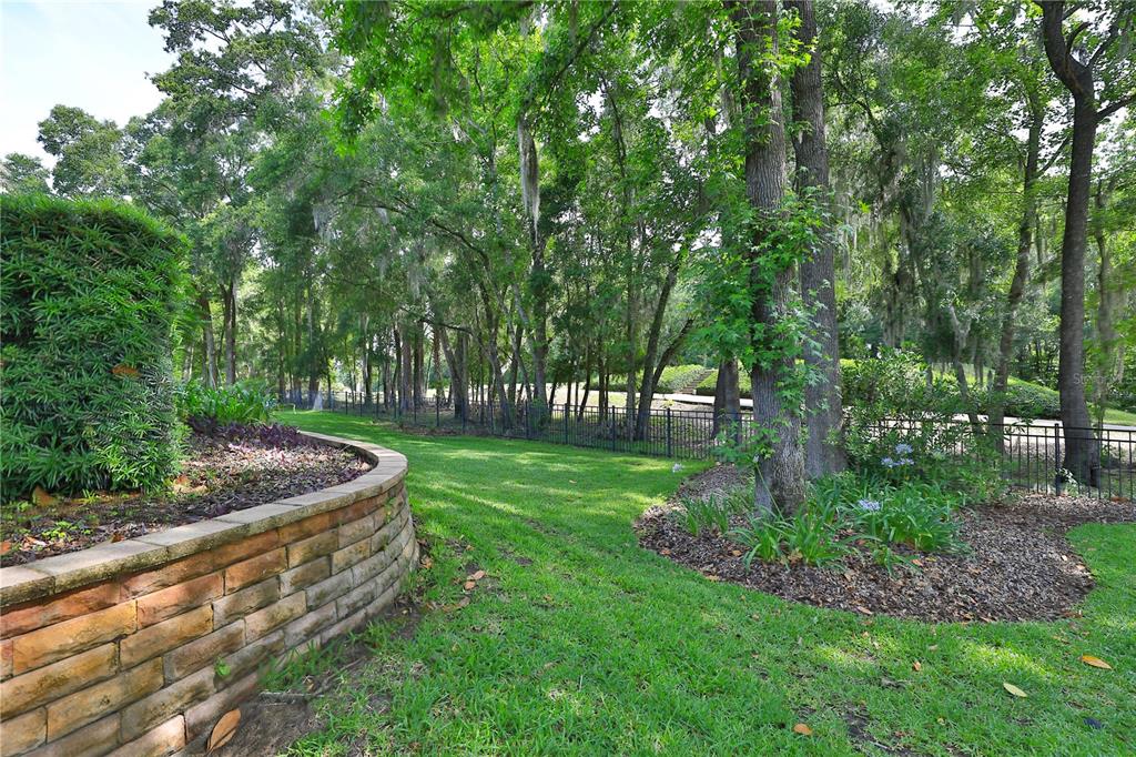 A curved brick retaining wall borders a grassy lawn with trees and a metal fence — 7122 SE 12TH CIRCLE, OCALA, FL
