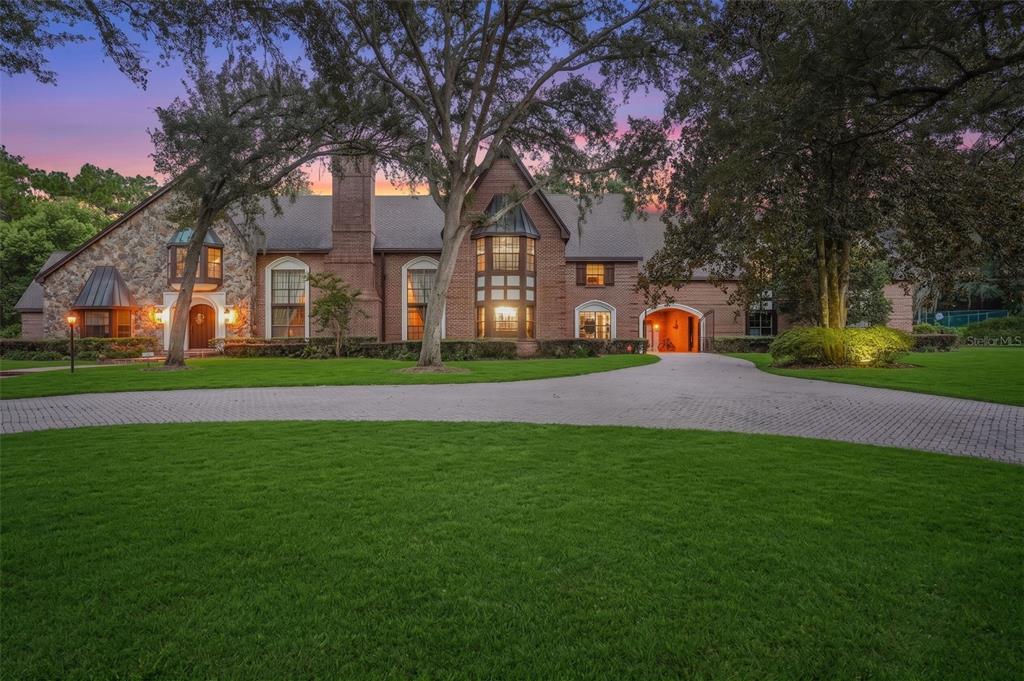 A large brick and stone house with a circular driveway and mature trees at dusk — 810 SWEETWATER CLUB BOULEVARD, LONGWOOD, FL