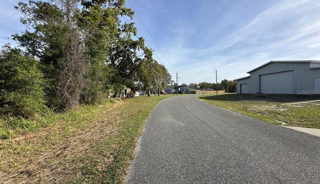 A paved road leads past a metal building and trees toward houses in the distance — 0 OLIVE CIRCLE, OCALA, FL