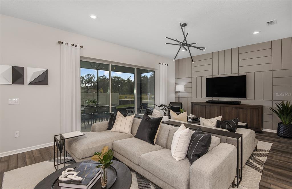 A modern living room features a sectional sofa, a geometric accent wall, and sliding glass doors — 5611 SYMPHONY SPRINGS TERRACE, PARRISH, FL