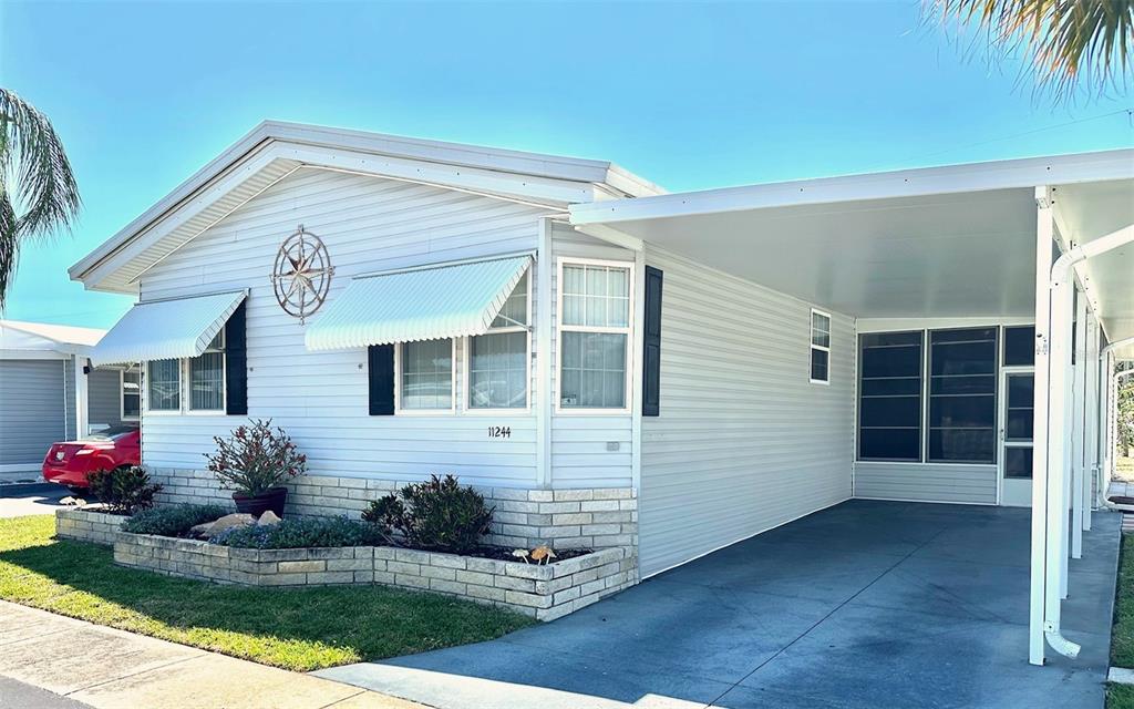 A light-colored manufactured home features a brick-accented planter, white siding, and an attached carport — 11244 101ST WAY UNIT 136, LARGO, FL