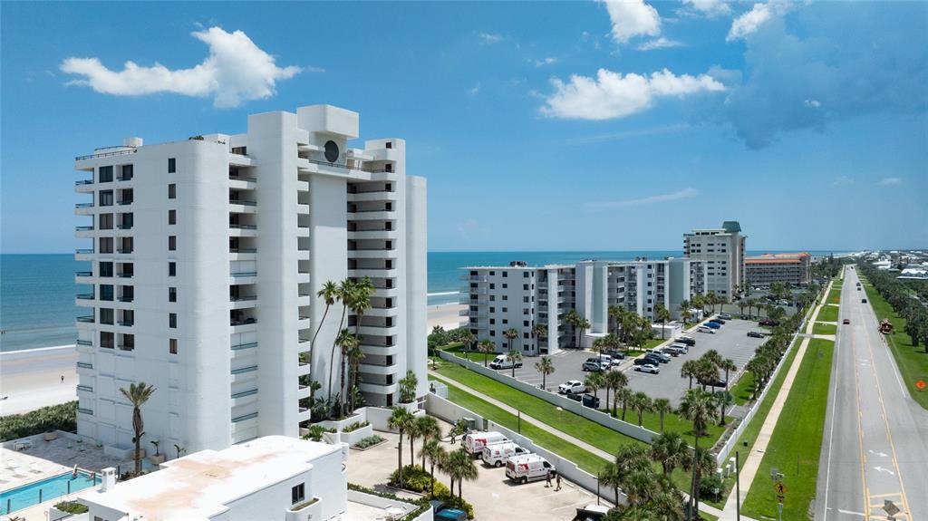 An aerial view shows white beachfront high-rise condominiums next to a road and parking lot — 5275 S ATLANTIC AVENUE UNIT 1007, NEW SMYRNA BEACH, FL