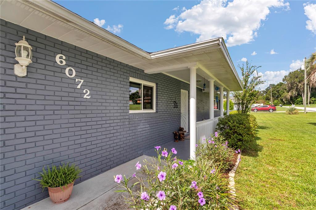 A gray brick house features a covered porch with white railings and purple flowers — 6072 E RUSH STREET, INVERNESS, FL