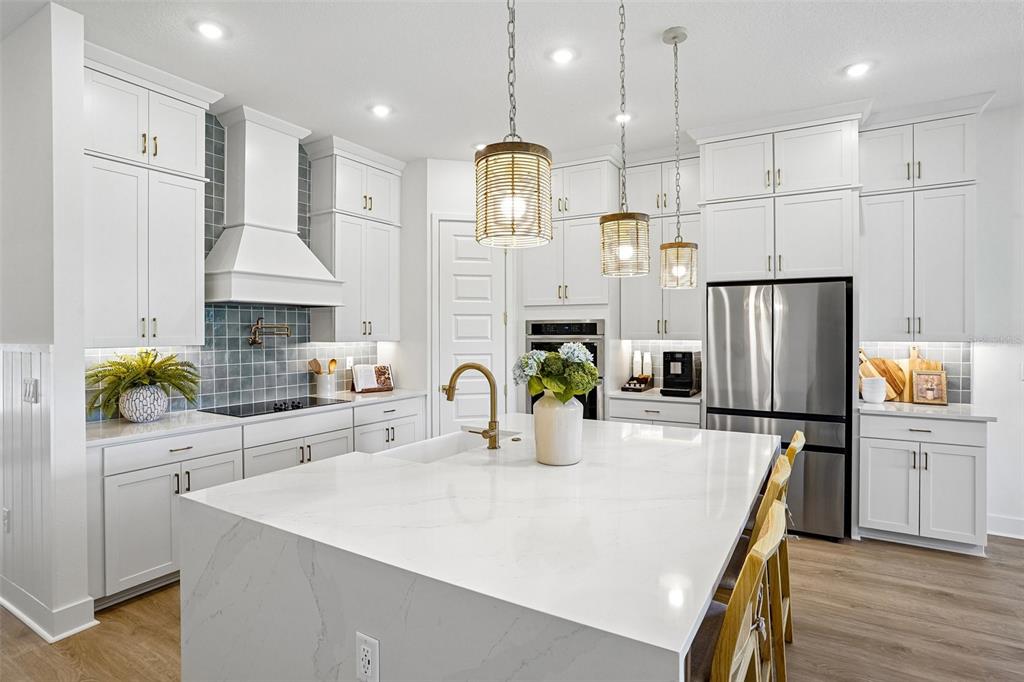 A modern white kitchen features a large quartz island, stainless steel appliances, and blue backsplash — 17428 HONEYBELL TANGELO COURT, WINTER GARDEN, FL