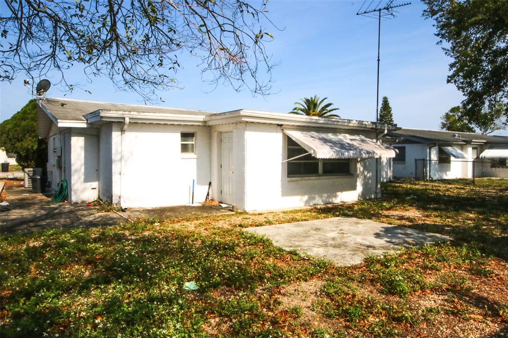 The exterior of a white, single-story house features a small concrete patio and metal awnings — 10824 QUEENS ROAD, PORT RICHEY, FL