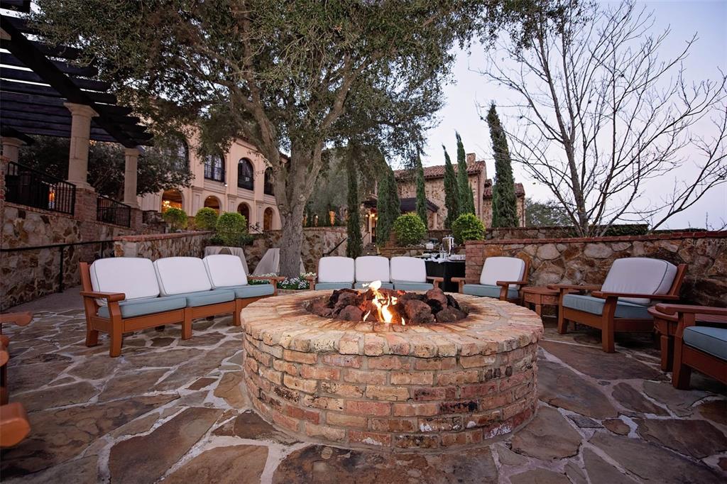 A stone patio features a circular brick fire pit surrounded by outdoor seating and trees — 16036 PENDIO DRIVE, BELLA COLLINA, FL